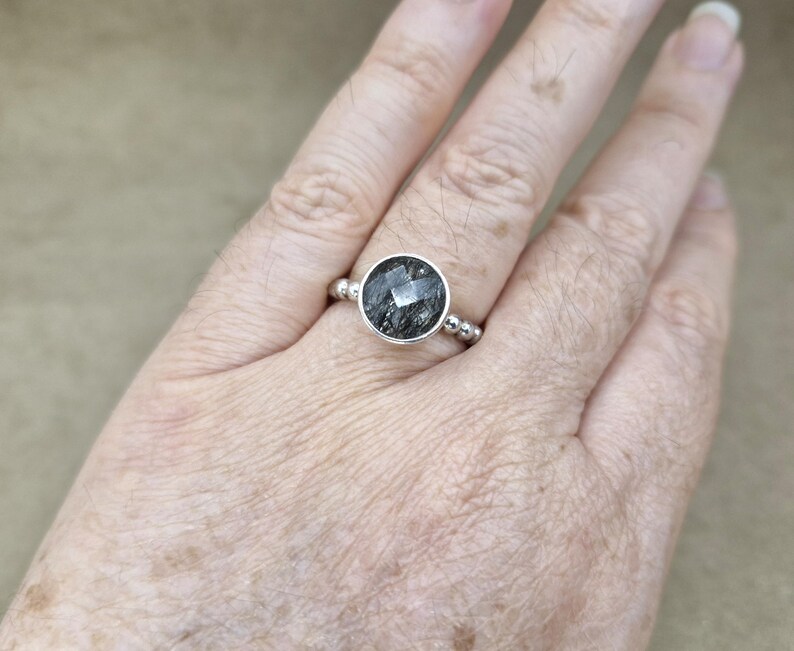 May include: A silver ring featuring a large, round, faceted black gemstone. The ring has a simple band with small silver beads on either side of the gemstone setting. The gemstone has a unique, textured appearance.
