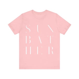 Puede incluir: Una camiseta rosa con el texto "SUNBATHER" impreso en blanco.