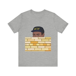 Può includere: Una maglietta grigia con un'illustrazione a cartone animato di una persona che indossa un berretto da baseball nero con le lettere "ATF" in giallo. La persona sta guardando oltre un muro di mattoni gialli.