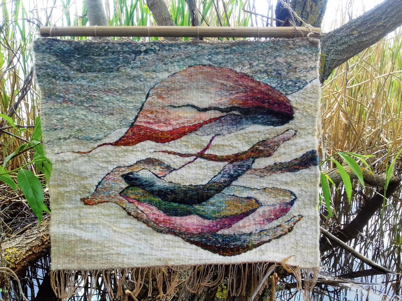 May include: A woven wall hanging with a cream background and abstract shapes in shades of blue, green, pink, and brown. The hanging has a fringe border.