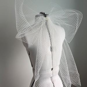 Puede incluir: Un velo de novia de tul blanco con un gran lazo en la espalda. El velo está drapeado sobre un maniquí blanco.