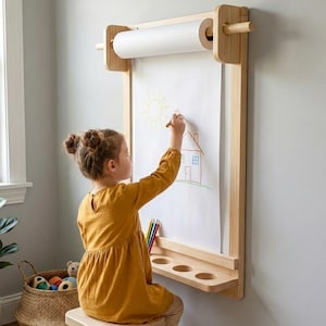 Peut inclure: Un chevalet mural en bois avec un rouleau de papier blanc et une étagère avec trois trous circulaires. Un enfant en robe jaune dessine sur le papier avec un crayon. Un panier de jouets est à proximité. L'enfant dessine une maison.