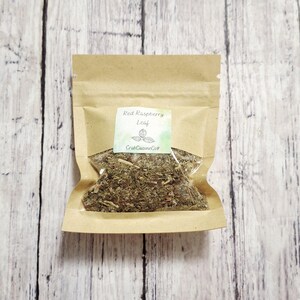May include: A small, brown paper bag filled with dried herbs, labeled "Red Raspberry Leaf." The label is a small rectangle with the text and a small illustration. The bag is on a light-colored, wooden surface.