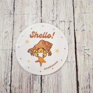 May include: A round sticker with a cartoon hermit crab wearing a shell, holding a starfish. The sticker has the text "Shello!" in brown, with small yellow stars around the edge. The sticker is on a wooden background.