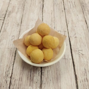 May include: A white bowl filled with small, round, yellow pastries on a white paper doily. The bowl is sitting on a light brown wooden surface.