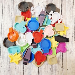 May include: A collection of colorful, 3D-printed shapes, including hearts, stars, skulls, and geometric forms. The assortment features various colors like red, blue, yellow, and pink. Some shapes appear to be small trays or containers.