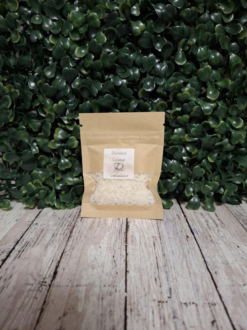 K&ouml;nnte beinhalten: Ein kleiner, rechteckiger Beutel aus braunem Papier mit einem durchsichtigen Sichtfenster, das wei&szlig;e Kokosraspeln enth&auml;lt. Ein wei&szlig;es Etikett auf der Vorderseite zeigt den Text "Shredded Coconut" &uuml;ber einer Kokosnuss-Grafik und "CrabCuisineCo".