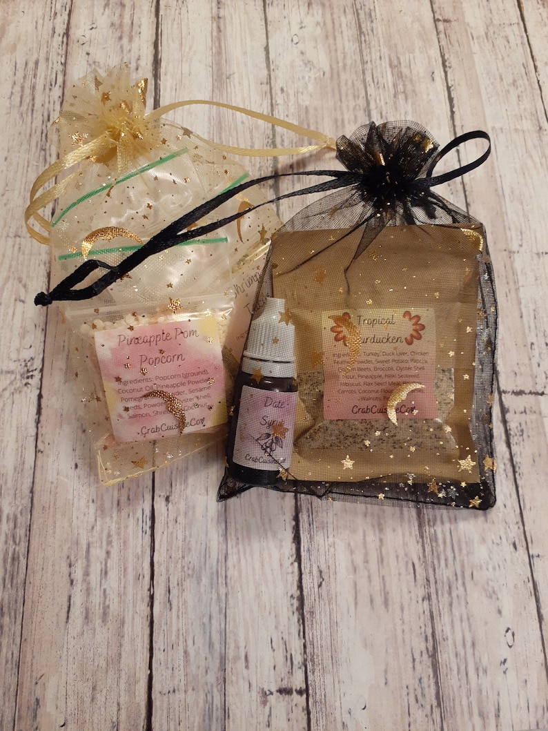 Puede incluir: Dos peque&ntilde;as bolsas llenas de palomitas de ma&iacute;z y una vela con aroma tropical. Las bolsas est&aacute;n hechas de tela transparente con estrellas doradas y est&aacute;n atadas con una cinta negra. La bolsa de palomitas de ma&iacute;z est&aacute; etiquetada como "Pineapple Pom Popcorn" y la bolsa de velas est&aacute; etiquetada como "Tropical Carducken".