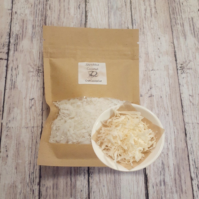 K&ouml;nnte beinhalten: Ein brauner Papierbeutel mit wei&szlig;er Kokosraspel und einem Etikett mit der Aufschrift Shredded Coconut und CrabCuisineCo. Eine kleine wei&szlig;e Sch&uuml;ssel, gef&uuml;llt mit denselben Kokosraspeln, steht vor dem Beutel auf einer Holzoberfl&auml;che.
