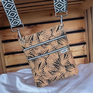 Könnte beinhalten: Eine Umhängetasche mit einem schwarz-weißen geometrischen Musterriemen. Die Tasche besteht aus einem korkähnlichen Material mit einem schwarzen Palmenblattmuster und drei Reißverschlussfächern. Sie hat silberfarbene Hardware und der Markenname "Ioanna" ist sichtbar.