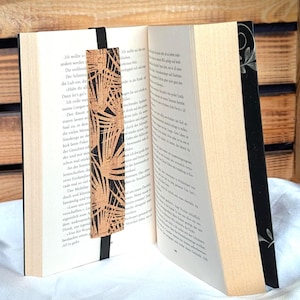Könnte beinhalten: Ein Buch mit einem dekorativen Lesezeichen. Das Lesezeichen ist aus Kork mit einem schwarzen Palmenblatt-Design. Das Buch hat einen schwarzen Buchrücken mit floralem Design und die Seiten sind cremefarben.