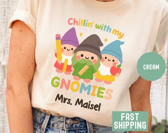 May include: Cream-coloured t-shirt with a colourful graphic of three cartoon gnomes. The gnomes hold a pencil, ruler, and apple. Text on the shirt reads "Chillin' with my GNOMIES Mrs. Maisel."