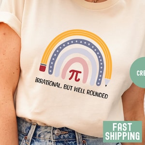 May include: Cream-colored t-shirt featuring a colorful rainbow design with a pencil on each side. The rainbow includes the pi symbol and the text "Irrational, but well rounded". The shirt is a casual wear item.