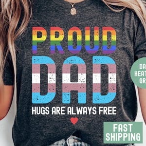 May include: A dark heather grey t-shirt with the text "Proud Dad" in rainbow and transgender pride colours. The text "Hugs are always free" is below the main text with a small red heart.