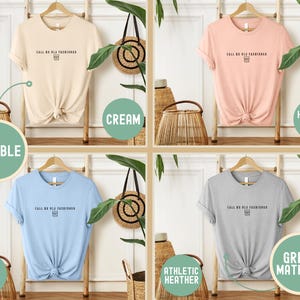 May include: Four t-shirts in cream, heather peach, baby blue, and athletic heather gray, each with the text "CALL ME OLD FASHIONED" and a small graphic. The shirts are displayed on wooden hangers.