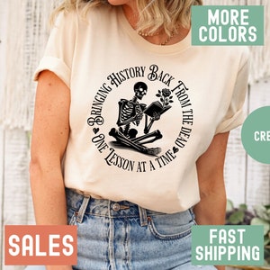 May include: A cream colored t-shirt with a black and white graphic of a skeleton reading a book. The graphic is in a circle with the text "Bringing History Back From The Dead One Lesson At A Time".