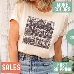 May include: A cream colored t-shirt with a brown graphic design of a farm scene. The graphic includes the text "Family Name Farms", "Smallville, AL, USA", "Est. 2024".