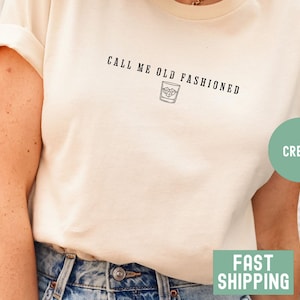 May include: Cream-colored t-shirt with the text "CALL ME OLD FASHIONED" above a graphic of a cocktail. The shirt has short sleeves and is paired with blue jeans. A green circle with the word "CREAM" is on the right side.