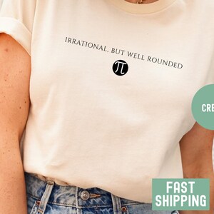 May include: Cream-colored t-shirt with the text "IRRATIONAL, BUT WELL ROUNDED" above a black Pi symbol. The shirt is made of a soft material and has rolled-up sleeves. The shirt is paired with blue denim jeans.