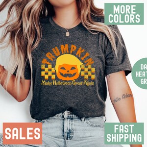 May include: A dark heather grey t-shirt with a pumpkin design featuring a face with a blonde wig and the text "TRUMPKIN" above the pumpkin and "Make Halloween Great Again" below the pumpkin.