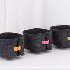 May include: Three grey felt storage baskets with different coloured leather handles. The handles are orange, pink and green.