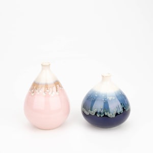 May include: Two ceramic vases with a unique glaze finish. The vase on the left is pink with a brown rim, while the vase on the right is blue with a white rim.