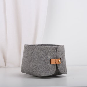May include: A gray felt storage basket with a brown leather handle and a gold metal buckle. The basket is sitting on a white surface.