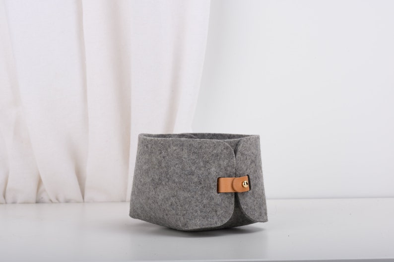 May include: A gray felt storage basket with a brown leather handle and a gold metal buckle. The basket is sitting on a white surface.