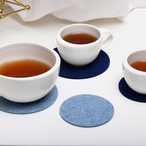 May include: Three white ceramic mugs with brown tea inside, sitting on blue and dark blue felt coasters.