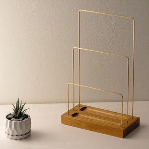 May include: A wooden desk organiser with three brass bars. The organiser has a rectangular wooden base with two holes and a small succulent plant in a geometric pot to the left. The brass bars are arranged in a tiered design.