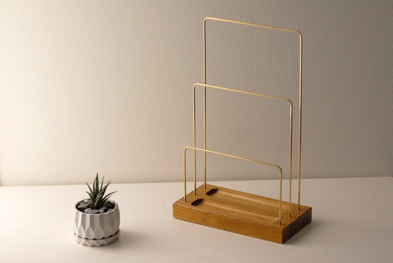 May include: A wooden desk organiser with three brass bars. The organiser has a rectangular wooden base with two holes and a small succulent plant in a geometric pot to the left. The brass bars are arranged in a tiered design.