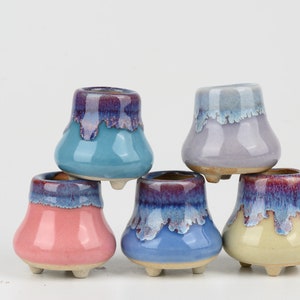 May include: Five small ceramic pots with a glazed finish in various colours, including blue, purple, pink, and yellow. Each pot has three small feet and a rounded shape.