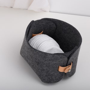 May include: A gray felt storage basket with a brown leather handle and a silver button closure. The basket is filled with white cotton rounds.