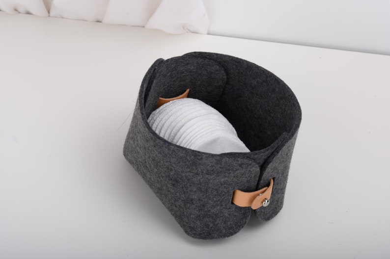 May include: A gray felt storage basket with a brown leather handle and a silver button closure. The basket is filled with white cotton rounds.