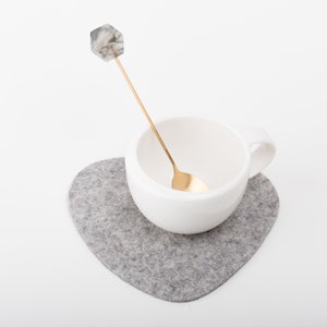 May include: A white ceramic mug with a gold spoon inside, sitting on a gray heart-shaped coaster.