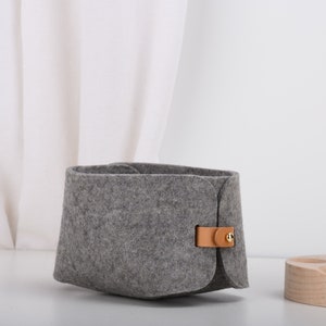 May include: A gray felt storage basket with a brown leather strap and a gold metal buckle. The basket is sitting on a white surface.