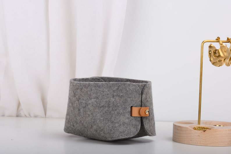 May include: A gray felt storage basket with a brown leather strap and a gold metal buckle. The basket is sitting on a white surface.