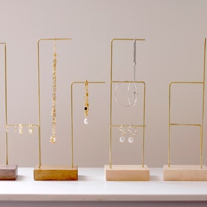 May include: Five gold metal jewelry stands with wooden bases. Each stand has a different configuration of bars for hanging earrings, necklaces, and bracelets. The stands are arranged in a row on a white surface.