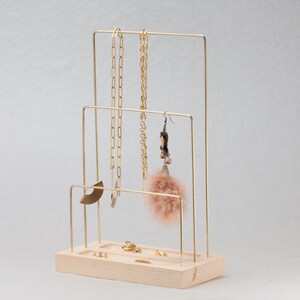 May include: A gold-coloured metal jewellery display stand with a light wood base. The stand has three levels for hanging necklaces and earrings. Several gold necklaces, earrings, and rings are displayed on the stand.