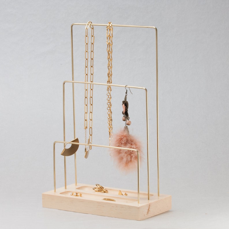 May include: A gold-coloured metal jewellery display stand with a light wood base. The stand has three levels for hanging necklaces and earrings. Several gold necklaces, earrings, and rings are displayed on the stand.