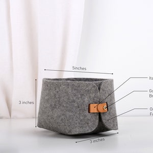 May include: A gray felt storage basket with a tan leather strap and brass hardware. The basket measures 5 inches wide and 3 inches tall.