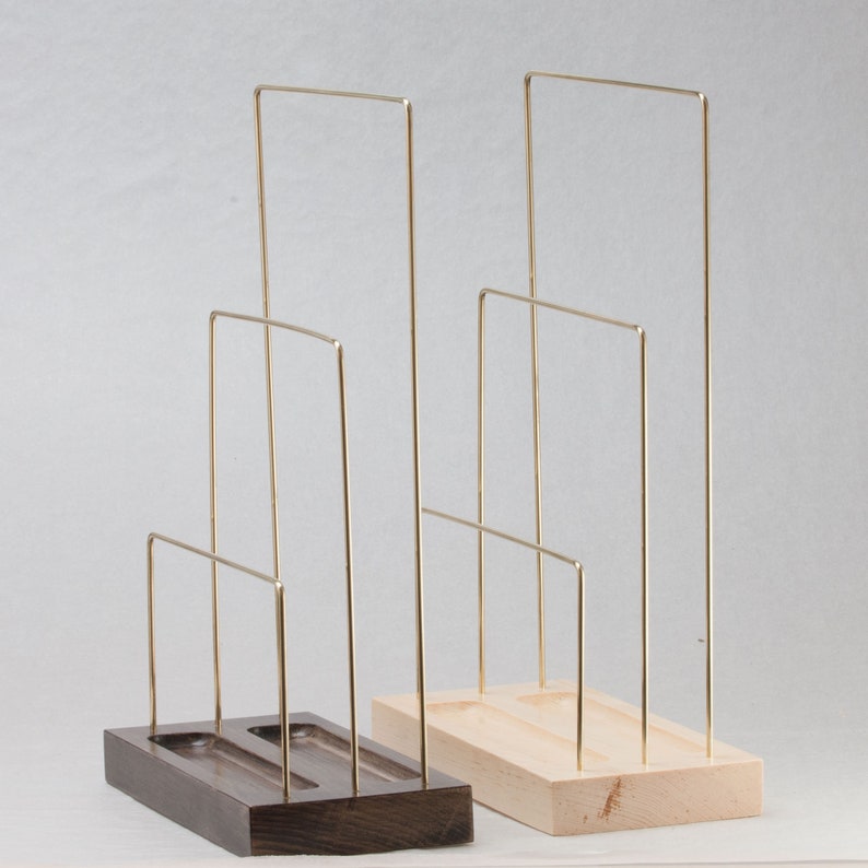 May include: Two jewellery display stands. One has a dark brown wooden base, the other a light wood base. Both have gold-coloured metal wire frames in a tiered design. The stands are designed to hold necklaces, bracelets, or other jewellery items.