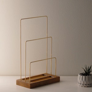 May include: A contemporary desk organiser with a wooden base and three gold-coloured metal dividers. The organiser is designed to hold papers or files. A small succulent plant in a geometric pot is nearby.