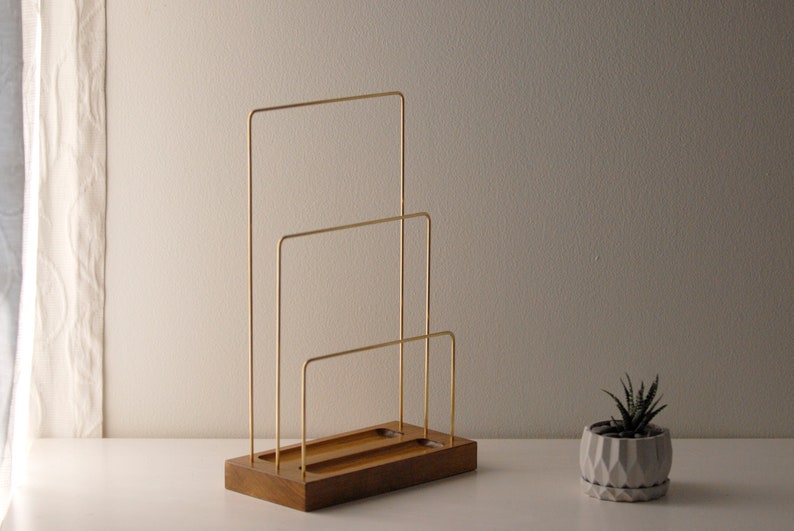 May include: A contemporary desk organiser with a wooden base and three gold-coloured metal dividers. The organiser is designed to hold papers or files. A small succulent plant in a geometric pot is nearby.