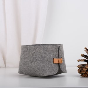 May include: A gray felt storage basket with a brown leather handle and a gold metal button. The basket is sitting on a white surface next to a brown pine cone.