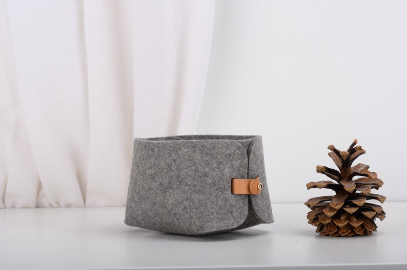 May include: A gray felt storage basket with a brown leather handle and a gold metal button. The basket is sitting on a white surface next to a brown pine cone.