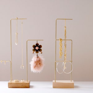 May include: Three gold metal jewelry stands with wooden bases. The stands are holding necklaces, earrings, and bracelets. A small gray velvet ring box is on the table.