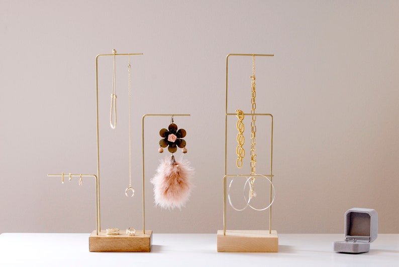 May include: Three gold metal jewelry stands with wooden bases. The stands are holding necklaces, earrings, and bracelets. A small gray velvet ring box is on the table.