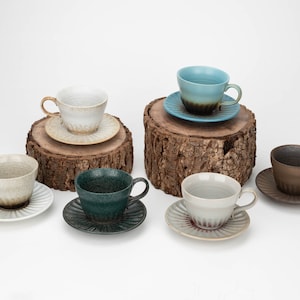 May include: A collection of ceramic teacups and saucers in various colors, including white, teal, and brown. The cups and saucers have a textured finish and are displayed on wooden blocks against a white background. The teacups have handles.