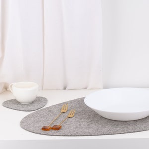 May include: Two gray felt placemats, one round and one oval, with a white ceramic bowl and a white ceramic cup on top. Two gold forks are placed on the oval placemat.